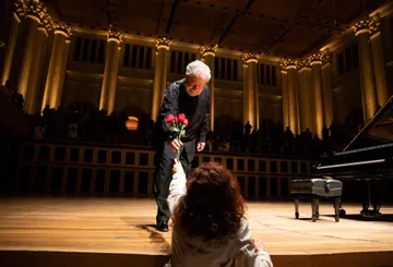 Foto de Nelson Freire, homem idoso, branco de cabelos curtos e brancos. Ele está em pé no palco da Sala São Paulo, ao lado de seu piano, recebendo rosas vermelhas de uma mulher que está no público, um nível abaixo do palco. Ele sorri.
