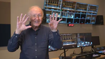 Foto de Heinz Holliger, homem idoso e branco, de cabelos ralos e brancos. Ele veste uma camisa de mangas longas, azul escuro e óculos retangular pequeno. Está com os dois braços levantados e as palmas das mãos viradas para a câmera, na altura da cabeça.