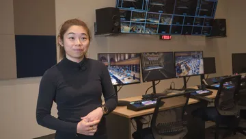 Foto de Elim Chan, mulher amarela, de pele clara, cabelos lisos e castanhos, presos num rabo de cavalo. Ela veste uma blusa preta, de manga longa e gol;a alta. Está parada em frente a diversas telas do estúdio de gravação da Sala São Paulo.