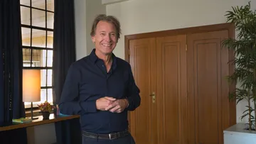 Foto de Marc Albrecht, homem branco e adulto, de cabelo castanho. Ele veste camisa de manga longa azul , calça jeans e cinto cinza. Eles sorri para a foto e segura uma mão com a outra, em frente ao corpo.
