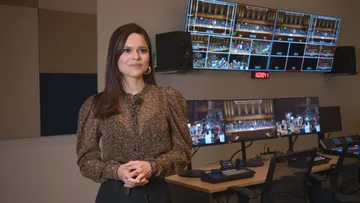 Foto de Larissa Martinez, mulher adulta, branca, de cabelos longos, lisos e castanhos. Ela veste uma blusa marrom com transparencia, abaixo dela uma regata preta. Ela está de pé, em frente a diversas telas do estúdio de gravação.