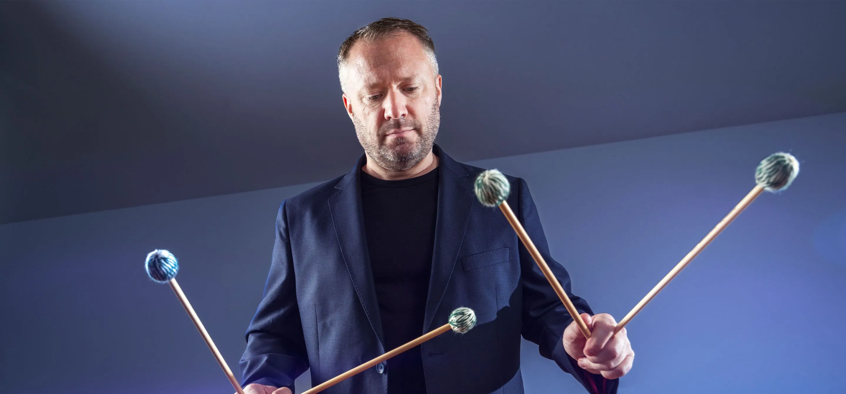 Colin Currie é um homem branco, na faixa dos 50 anos. Ele usa um paletó preto e segura nas mãos baquetas de percussão.