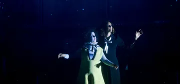 A imagem mostra uma palhaça de roupa amarela e a regente Alondra de la Parra, uma mulher branca de cabelos castanhos, em uma caixa preta.