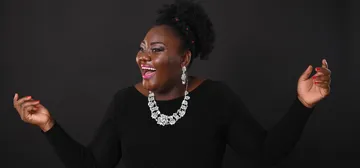 Masabane é uma mulher negra, de cabelos pretos presos em um coque. Ela usa batom rosa, colar e brinco brilhantes e vestido preto.