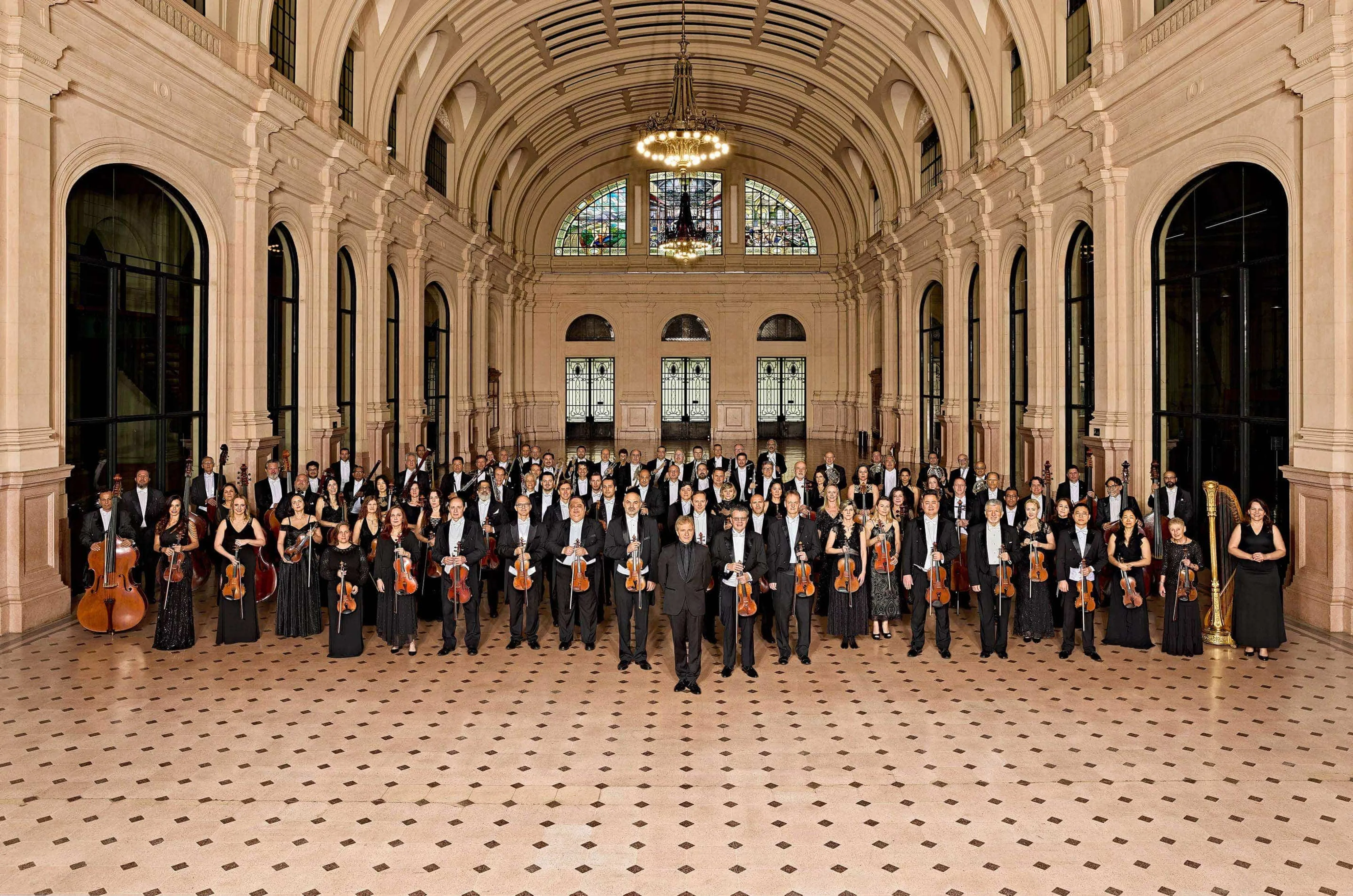 Os músicos da Orquestra estão na Estação das Artes, um grande salão com vitrais e piso cor de rosa. Eles vestem preto, seguram seus instrumentos e sorriem.
