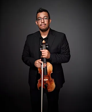 Leandro Dias é um homem negro, de cabelos castanhos e óculos. Ele está vestindo uma camisa preta e um blazer.