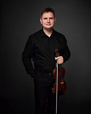 Alexey Chashnikov is a white man with brown hair. He is wearing a black shirt and holding a violin.