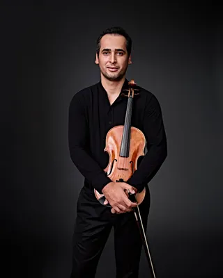 Foto de André Rodrigues, um homem branco, de cabelos pretos e curtos. Ele está usando uma camisa social preta com o primeiro botão da gola aberto. Segura sua viola à frente do peito.