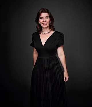 Clarissa é uma mulher branca, com cabelo castanho e olhos castanhos. Ela está usando um vestido preto e um colar com pedra preciosa.