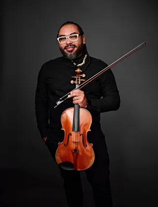 Ederson Fernandes é um homem negro, com tranças, barba e óculos brancos. Ele está usando uma camisa preta e segurando uma viola.