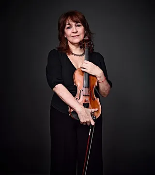 Elina Suris is a white woman with brown hair and bangs. She is wearing a low-cut black dress and a stone necklace, and she is holding a violin.
