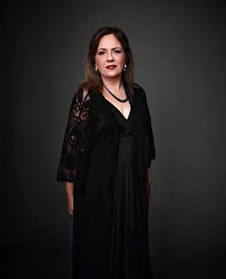 Lea Lacerda é uma mulher branca, com cabelo loiro e longo. Ela está usando um vestido preto e um colar.