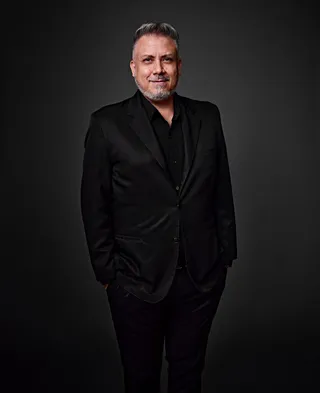 Luiz Guimarães é um homem branco, com cabelo grisalho, barba e bigode. Ele está usando uma camisa preta e um paletó preto.