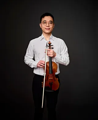 Photo of Marcio Kim. He is a light-skinned Asian man with short black hair. He is wearing a white dress shirt and round glasses. He is holding his violin in front of his chest.