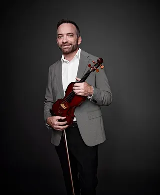 Matthew Thorpe é um homem branco, de olhos claros, cabelos castanhos e barba. Ele está usando uma camisa branca e um paletó cinza, e segurando um violino.