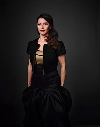 Natália Áurea é uma mulher branca, com olhos claros. Ela tem cabelo escuro e longo, preso de lado. Está usando um vestido dourado com um bolero preto.