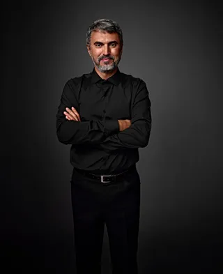 Odorico é um homem de pele média, com cabelo grisalho, barba e bigode. Ele está usando uma camisa preta e está com os braços cruzados. Há uma pinta marrom abaixo do olho direito.