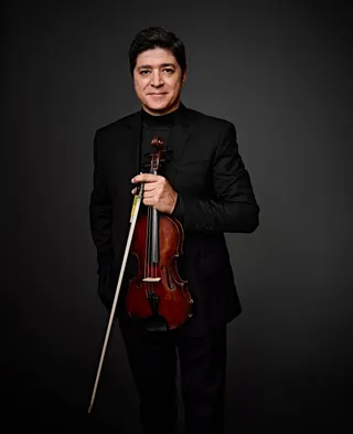 Paulo Paschoal is a white man with brown hair. He is wearing a black shirt and suit jacket, and he is holding a violin.